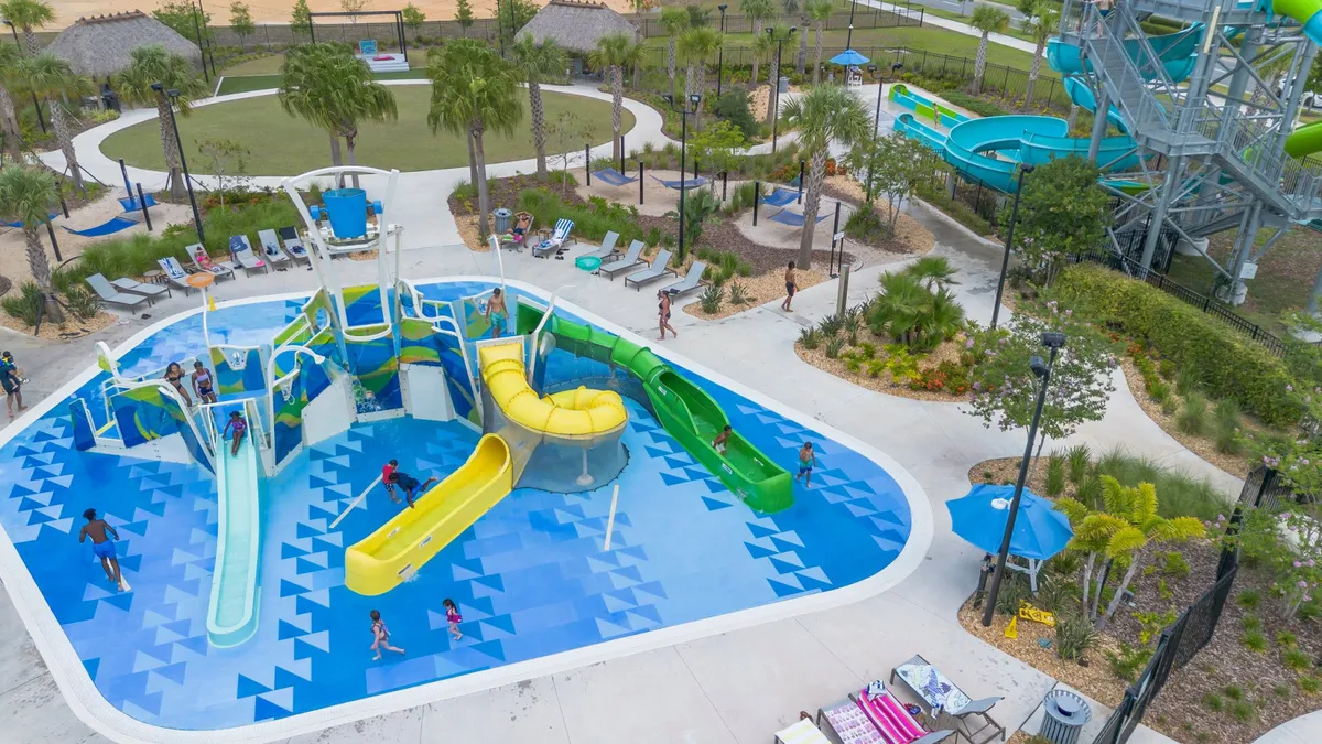 A joyful exterior view of a pool complex filled with children playing in the water, colorful water slides, lounge chairs for relaxation, and well-maintained landscaping. The scene conveys energy and excitement, with families enjoying the water park environment.