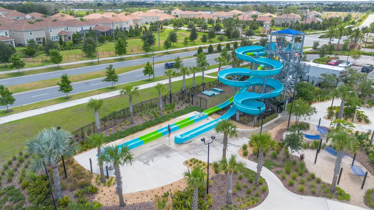 A lively water play area featuring a colorful water slide, surrounded by palm trees and landscaped greenery. Families and children are shown enjoying the water features and sunbathing in the lush communal areas, emphasizing energetic, playful community experiences.