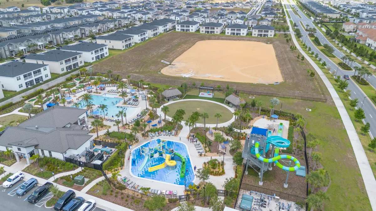 A vibrant exterior scene showing a large swimming pool with water slides, green play areas, and surrounding community buildings. Children are playing, families are gathered around, and the area is lively and inviting for all ages.