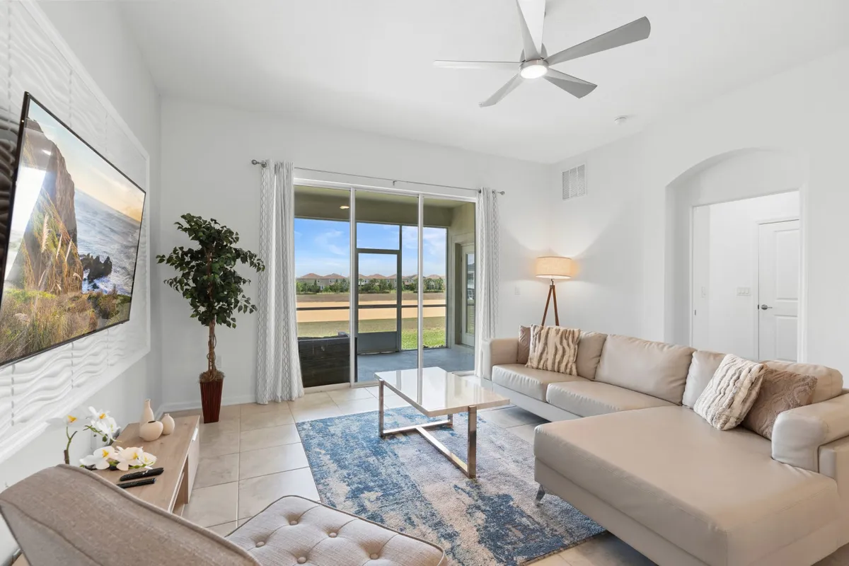 The image showcases a bright and spacious living room with a plush sofa set arranged around a modern coffee table. Floor-to-ceiling windows bathe the space in natural light, offering sweeping sunset views. A large television with a scenic image, potted plant, and comfortable seating create a serene setting perfect for relaxation or gathering with loved ones.