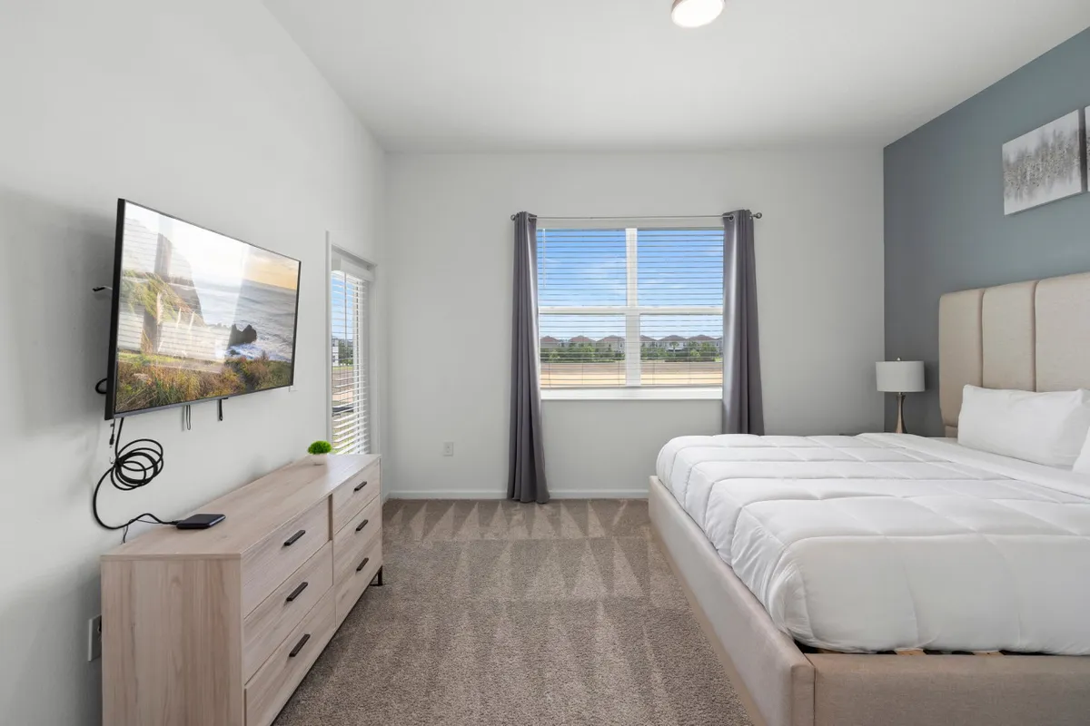 A cozy bedroom featuring a bed, a Smart TV, dresser, and a window with a soothing view. The setup invites guests to unwind with evening shows or enjoy the calming scenery.