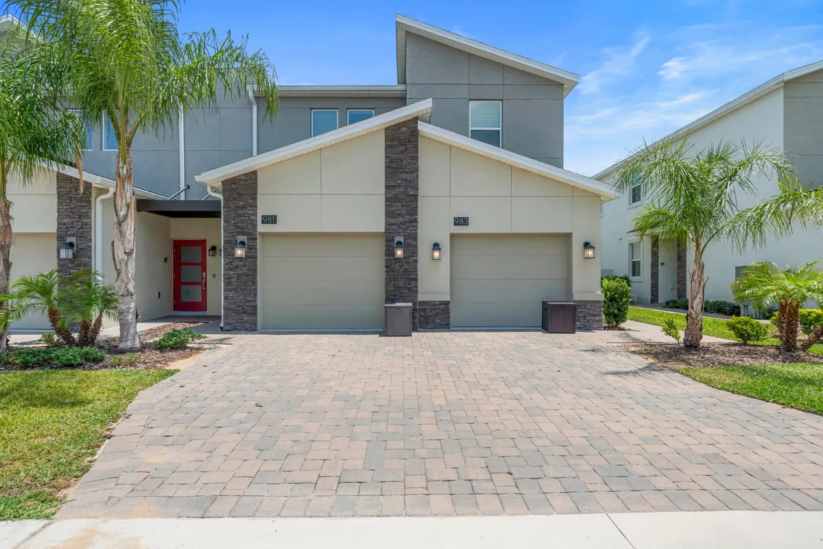 A welcoming exterior scene featuring modern architecture, a paved driveway, landscaped garden, palm trees, and a vibrant red front door. The setting conveys a sense of arrival, with space suitable for outdoor gatherings or welcoming friends.