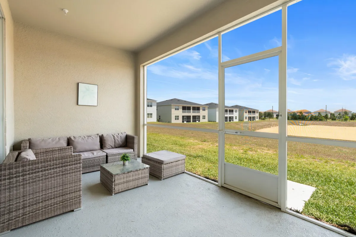 A cozy indoor patio seating area with wicker chairs, a small coffee table, and large windows that open onto a green lawn. The natural light, greenery, and comfortable arrangement make it an inviting spot for relaxing with a morning coffee or enjoying evening breezes.
