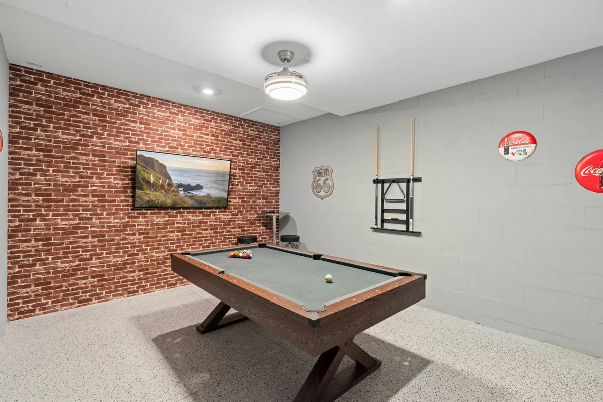 Another angle of the game room, this image features the pool table, a brick accent wall, television displaying landscape art, and Coca-Cola memorabilia, providing additional context of the space’s layout and entertainment options.